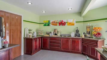 Una cocina con armarios de madera, una encimera con diversos electrodomésticos y alimentos, y una pared decorada con imágenes de frutas y verduras.