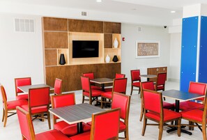 A modern dining area with red upholstered chairs and wooden tables, a flat-screen TV mounted on a wooden panel, and a blue storage unit in the background. - Holiday Inn Express Greenville by IHG (Greenville)