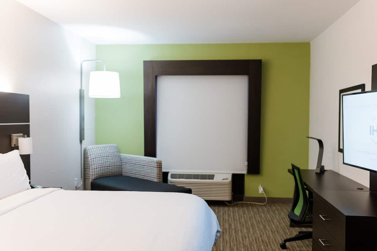A hotel room with a bed, a desk, a chair, a projector screen, and an air conditioning unit.