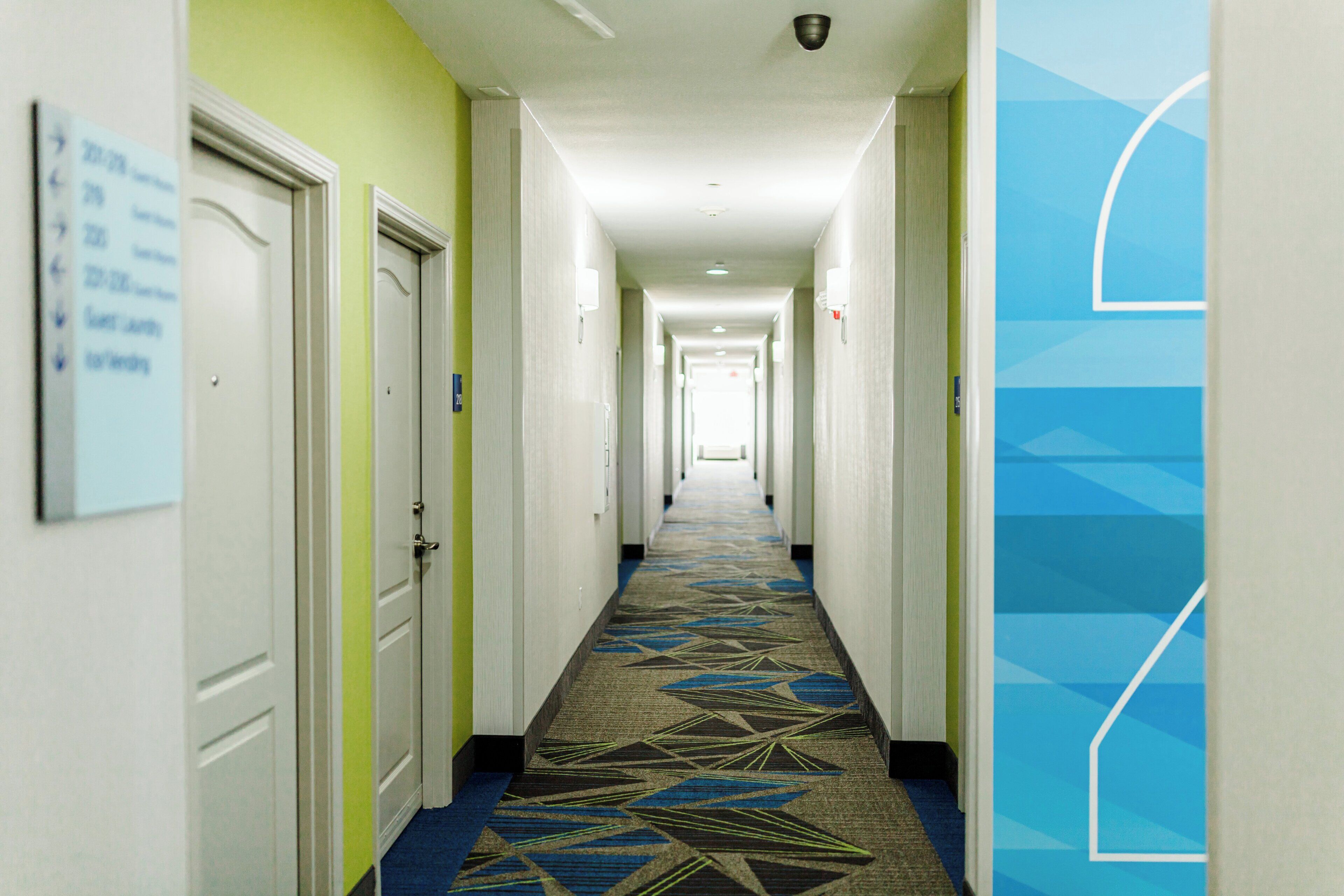 Pasillo de hotel con puertas a ambos lados, una alfombra estampada y un diseño geométrico azul en la pared.