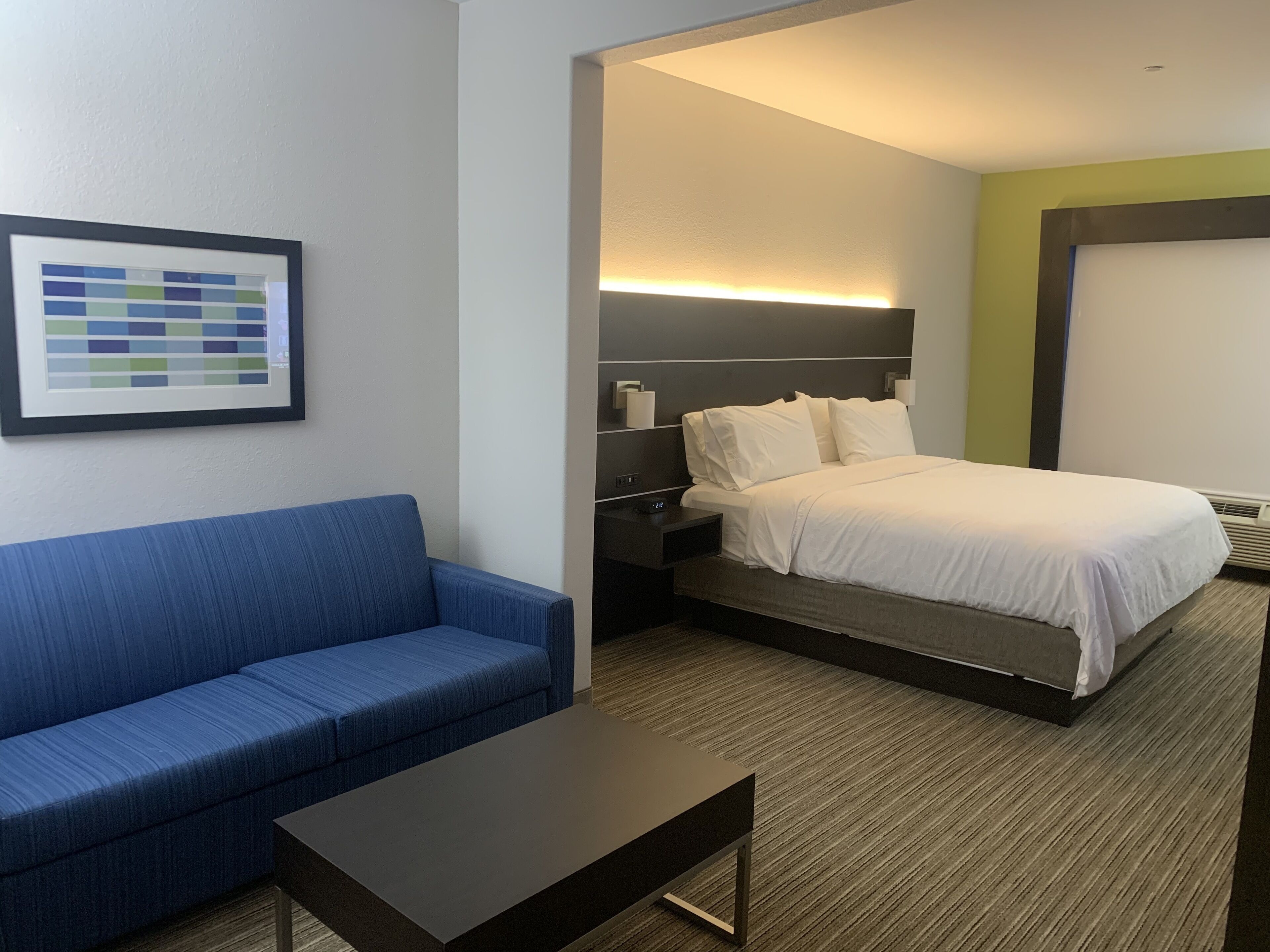 A hotel room with a blue sofa, a small table, a bed with white bedding, a nightstand, and a wall-mounted light.