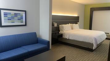 A hotel room with a blue sofa, a small table, a bed with white bedding, a nightstand, and a wall-mounted light.