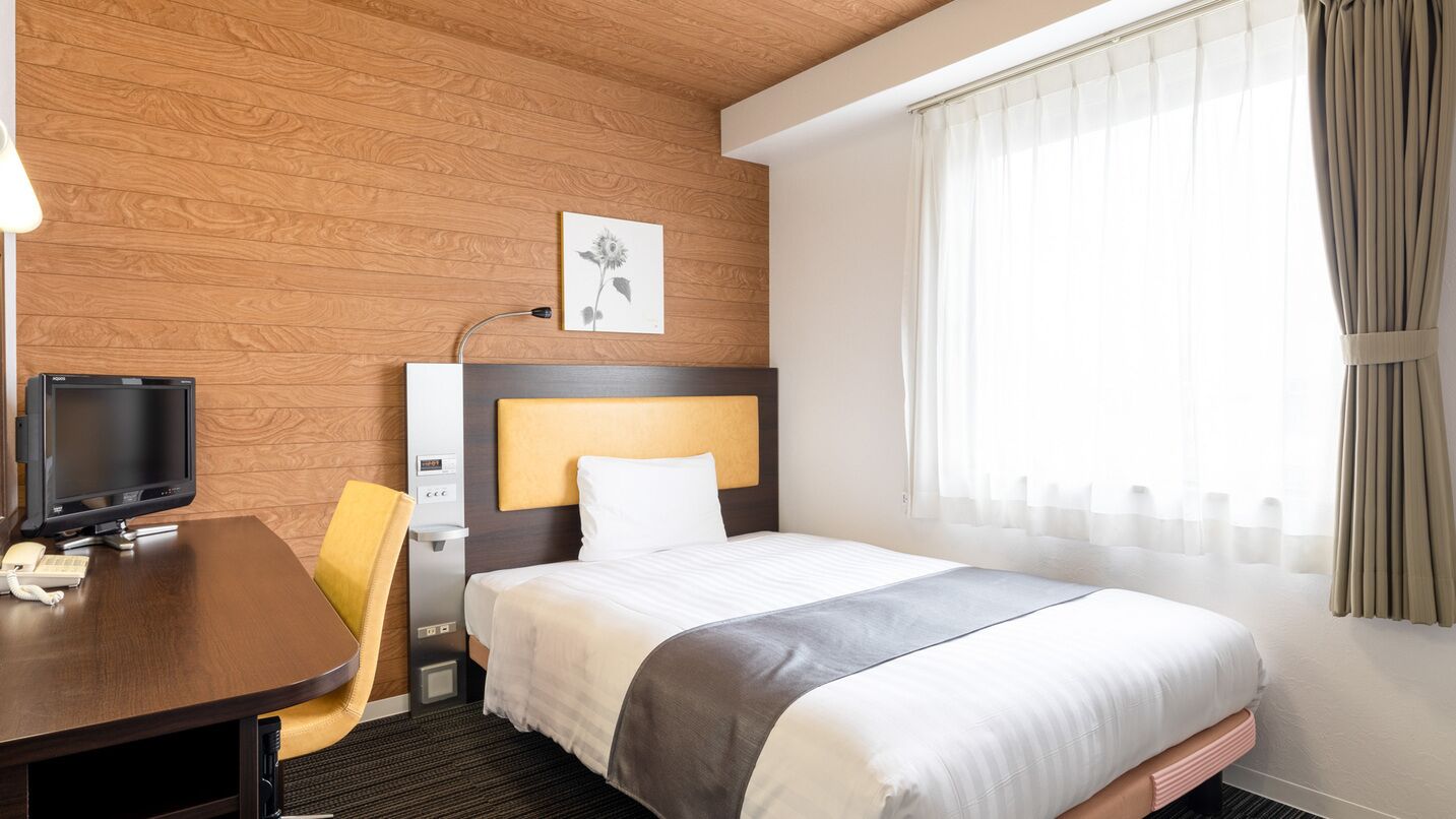 A hotel room with a wooden wall, a bed with a white headboard, a desk with a computer and phone, and a window with curtains.