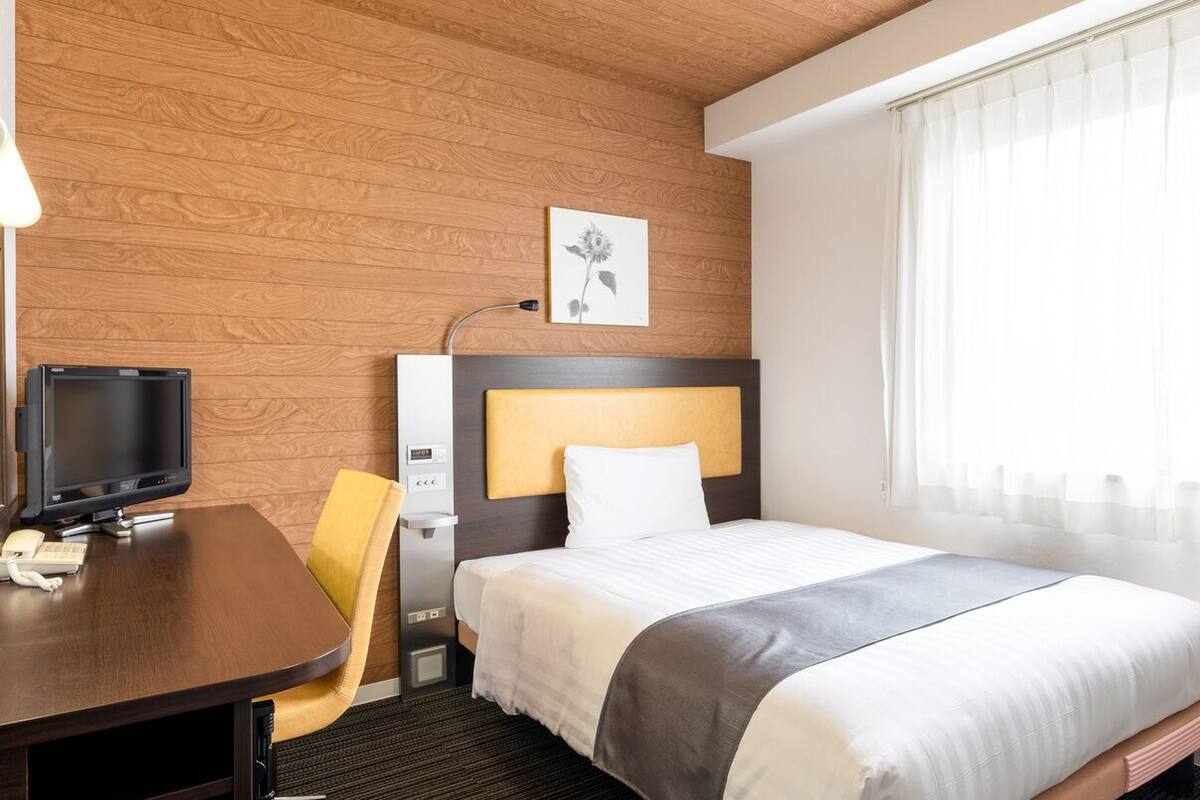 A hotel room with a wooden wall, a bed with a white headboard, a desk with a computer and phone, and a window with curtains.