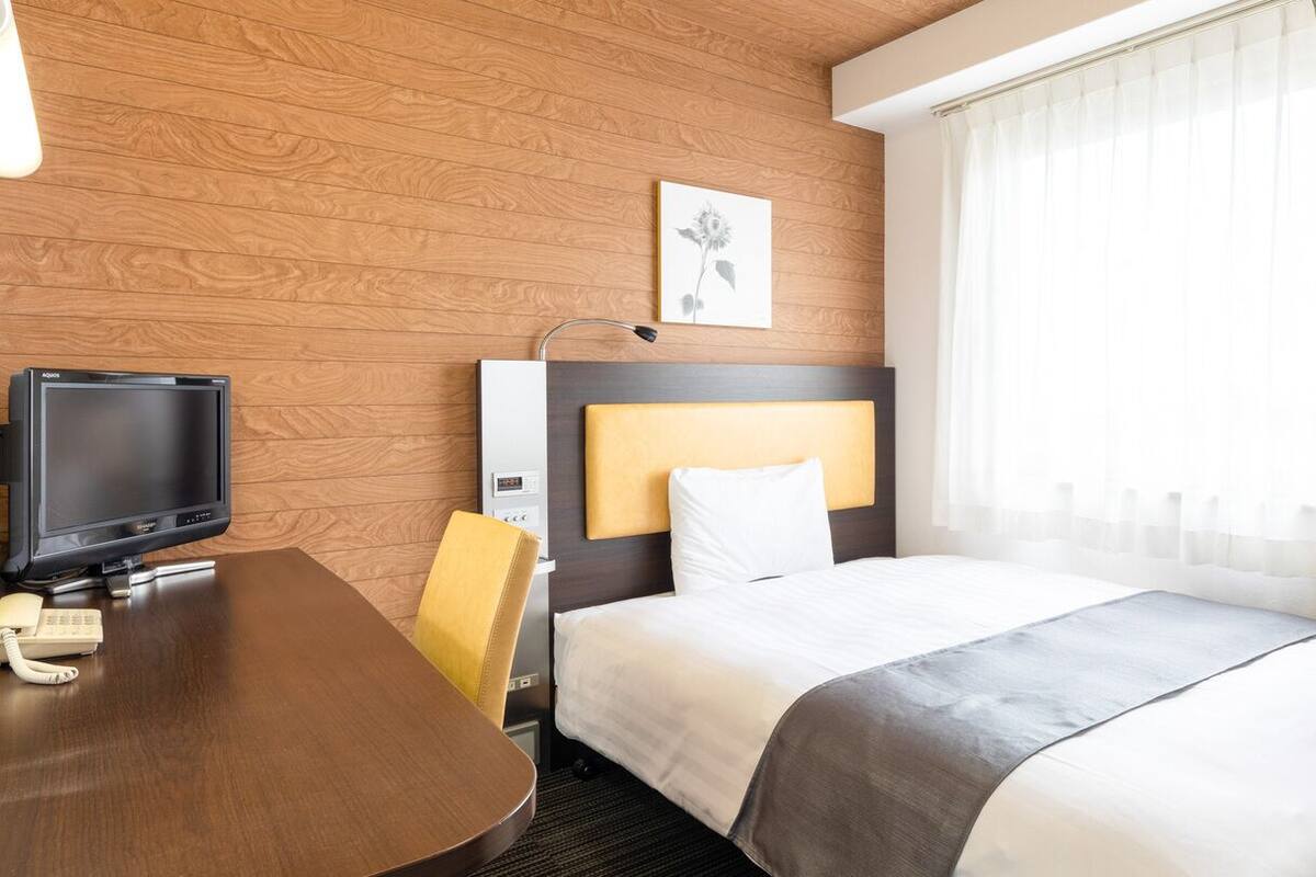 A hotel room with a wooden wall, a bed with white bedding, a desk with a television, and a telephone.