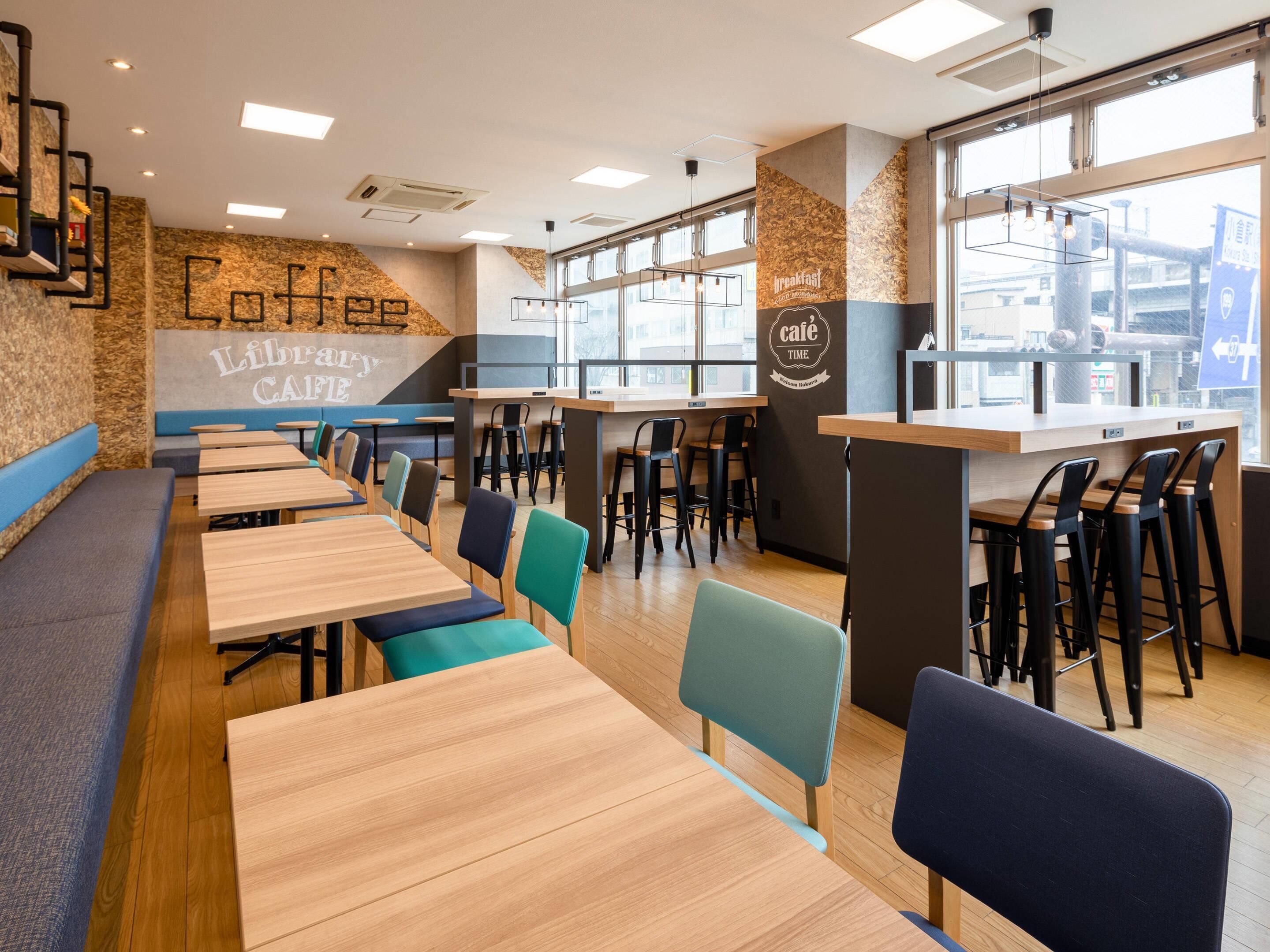 A café with long wooden tables and colorful chairs, a counter with stools, and a wall with the words "Coffee Library Café".
