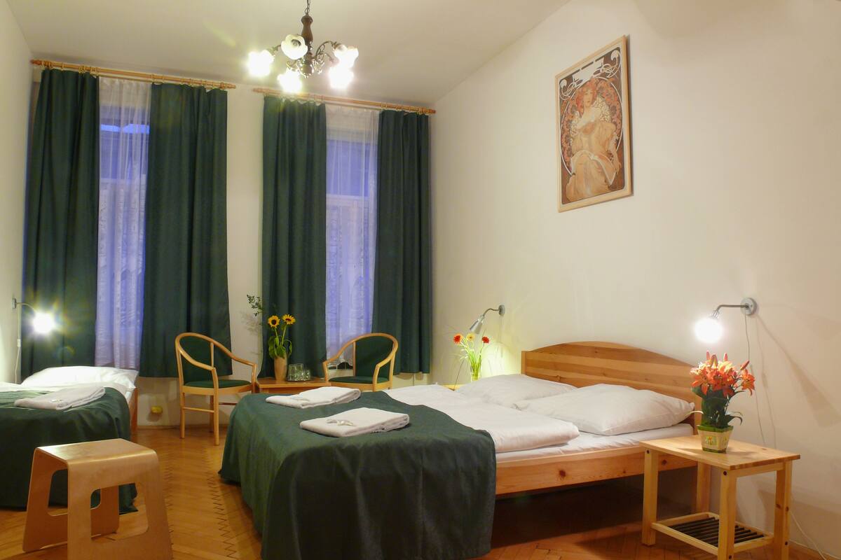 A hotel room with two beds, a wooden headboard, a nightstand with a lamp, a small table with a vase of flowers, and a painting on the wall.