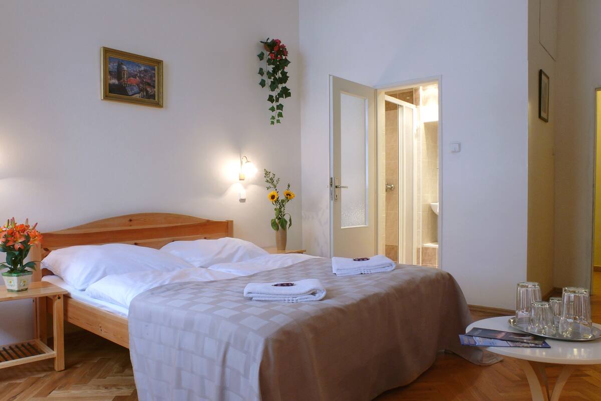 A neatly made bed with white linens, a bedside table with a vase of flowers, a wall-mounted picture, and a bathroom door visible in the background.