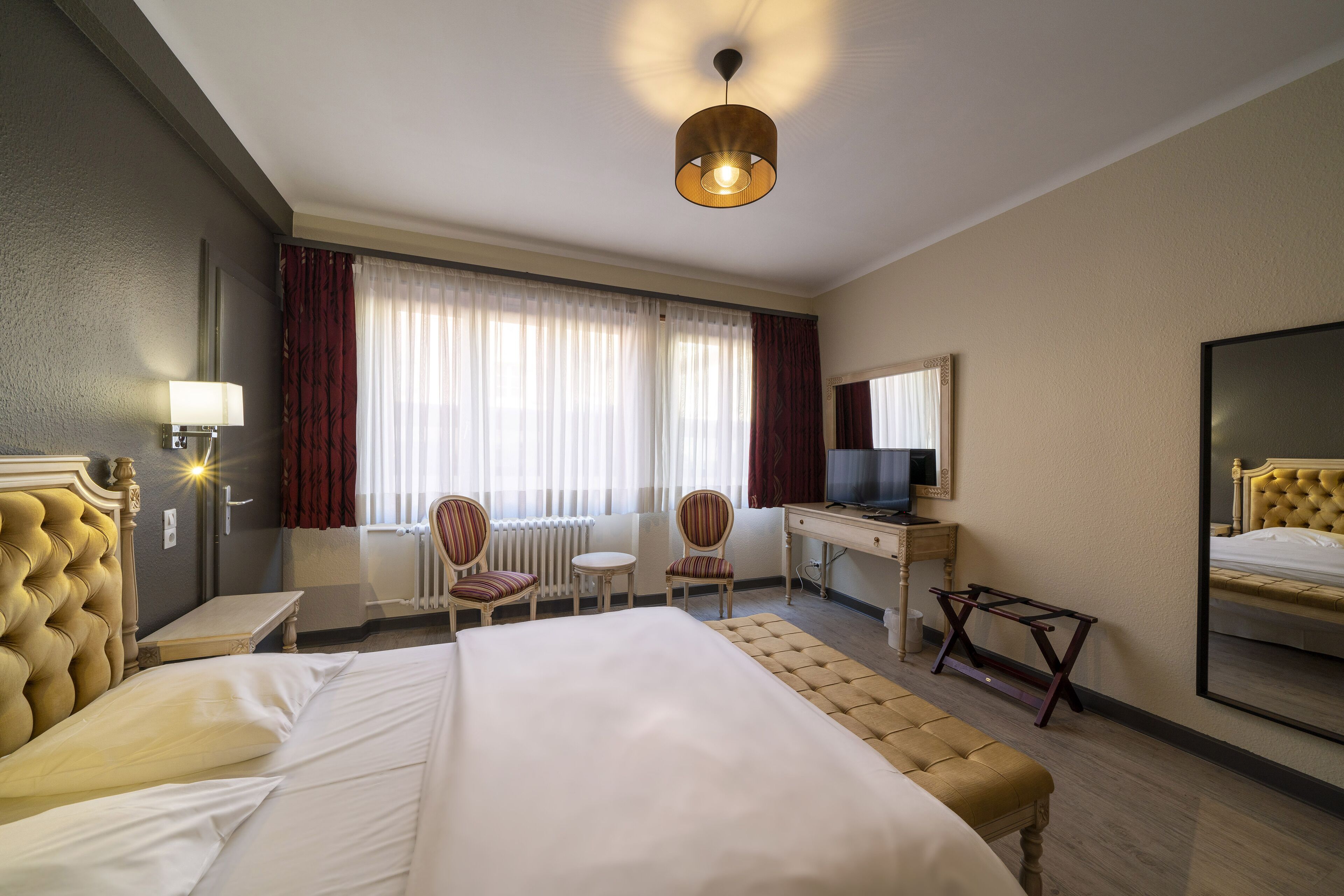 Ein Hotelzimmer mit einem großen Bett, zwei Stühlen, einem Schreibtisch mit Computer, einem Fernseher und einem großen Spiegel.
