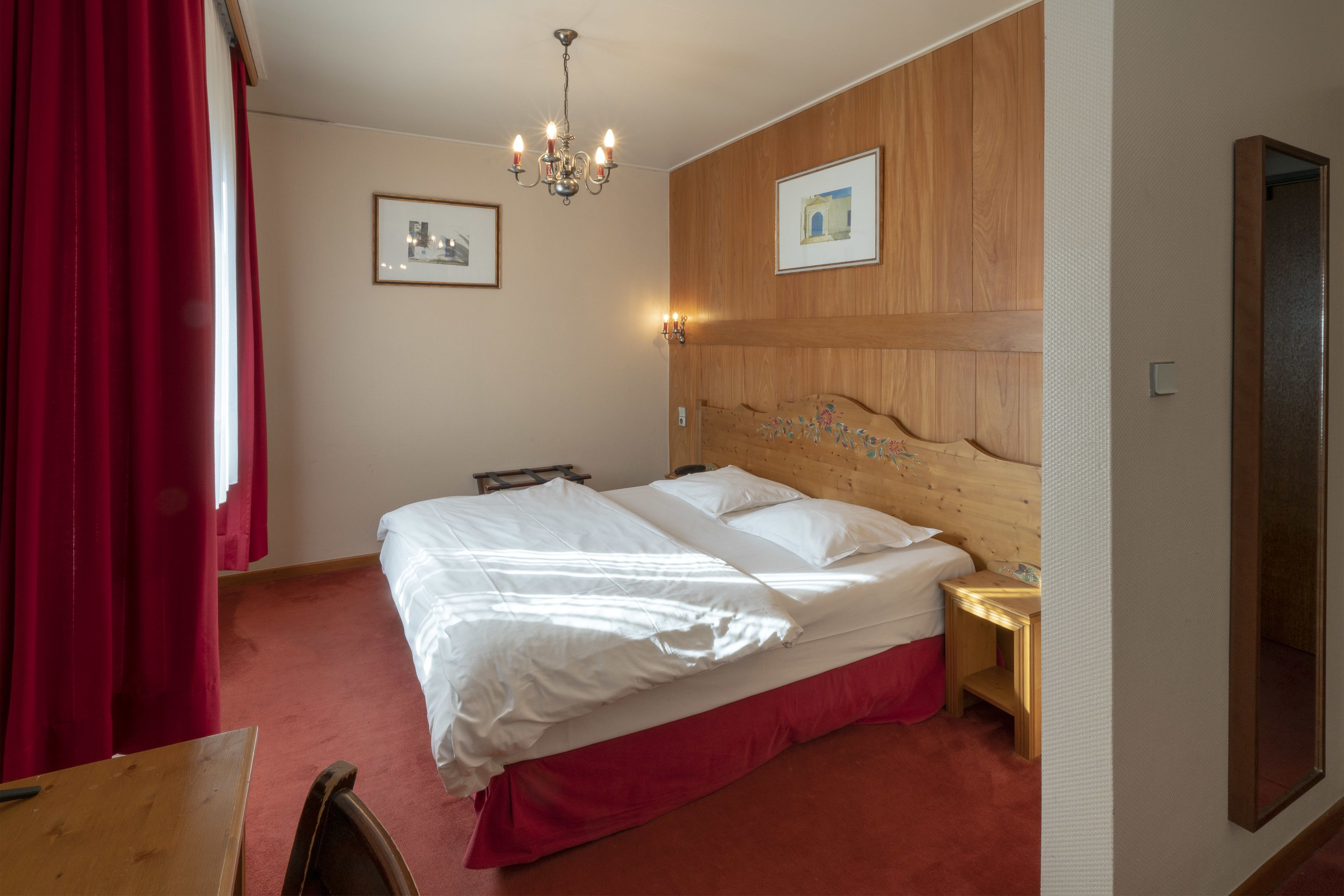 A hotel room with a wooden headboard, a large bed with white linens, a bedside table with a lamp, and a red curtain.