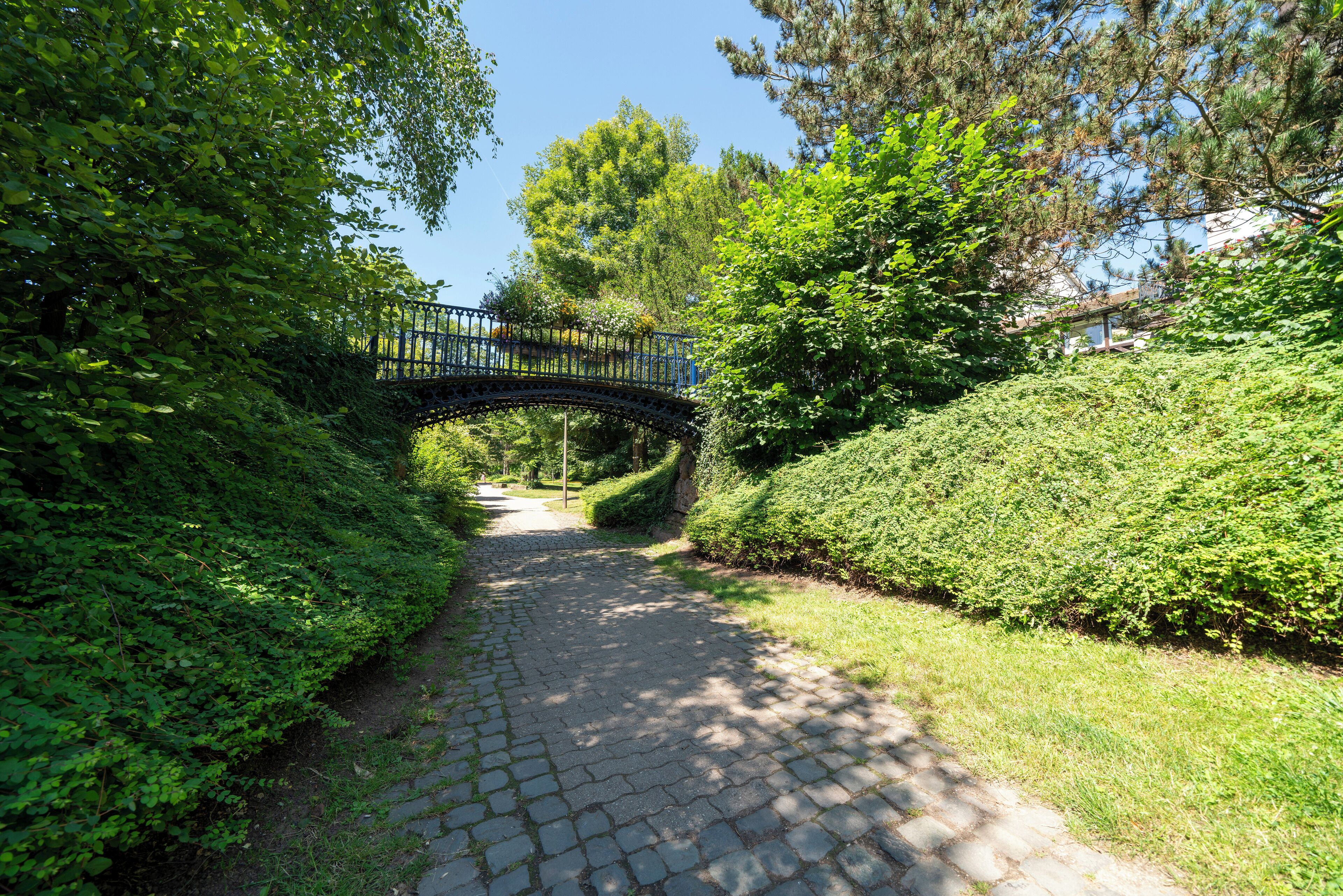 A cobblestone path leading through a park with a metal bridge over it.
