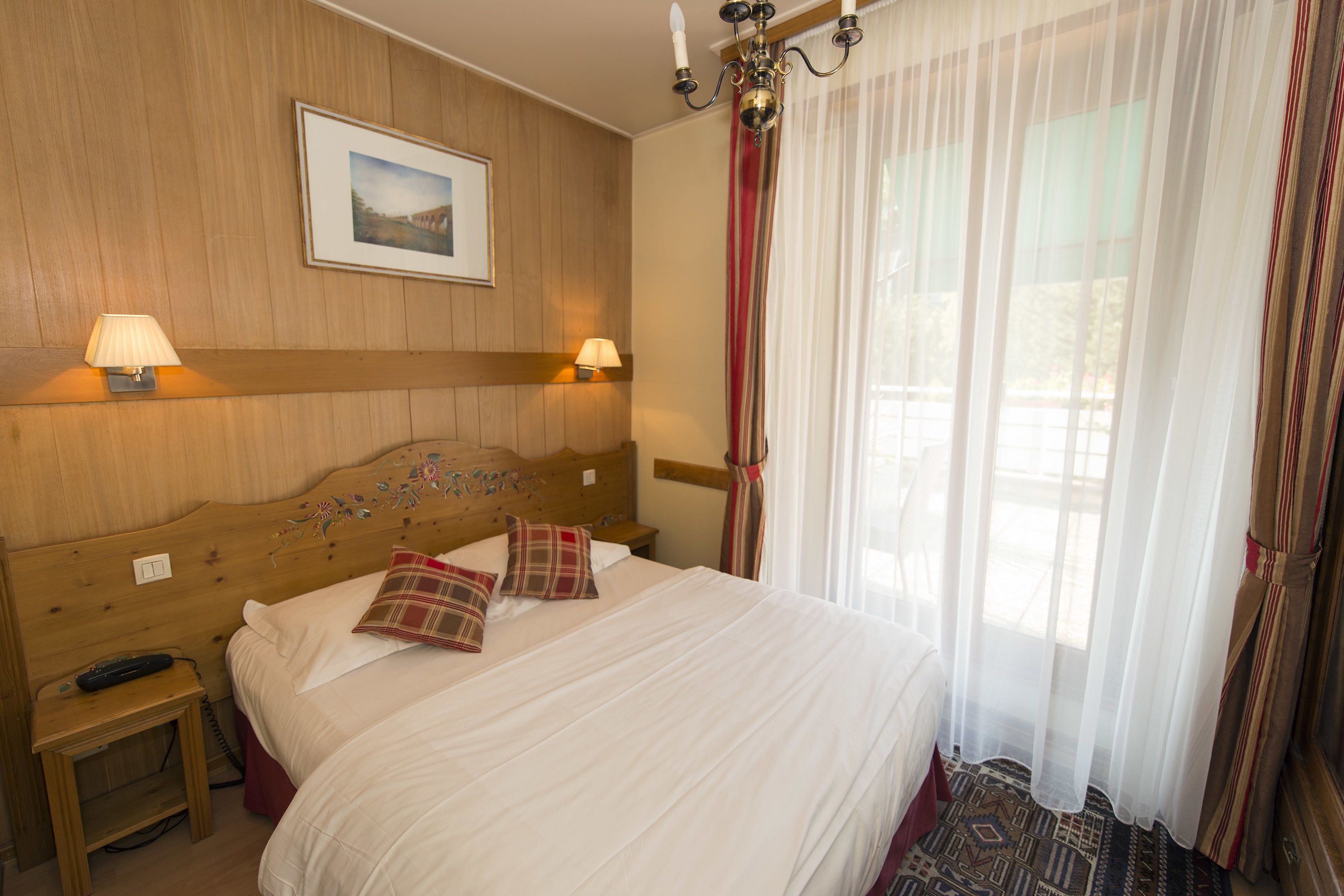 Habitación de hotel con cabecera de madera, dos lámparas de noche, cama con ropa de cama blanca y almohadas escocesas, una mesita de noche con un control remoto y un ventanal con cortinas traslúcidas.