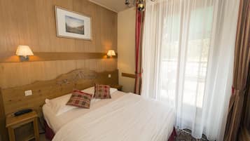 A hotel room with a wooden headboard, two bedside lamps, a bed with white linens and plaid pillows, a small bedside table with a remote control, and a large window with sheer curtains.