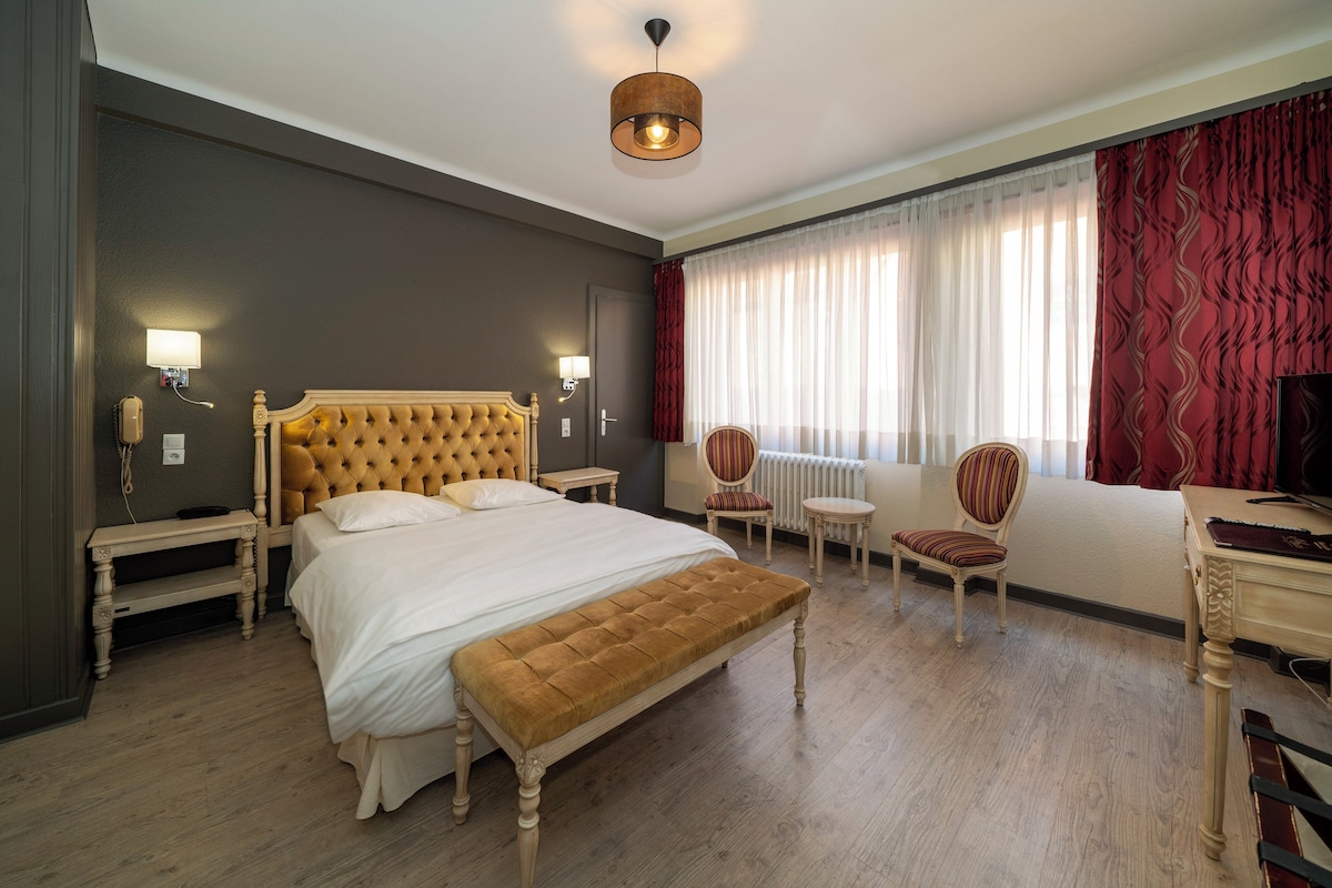 A hotel room with a large bed, a bench, a chair, a small table, and a television.