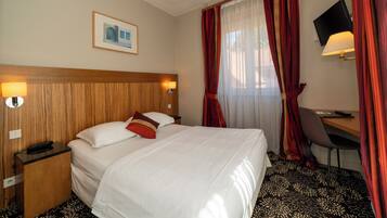 A hotel room with a large bed, a desk with a chair, a TV, and a window with red curtains.