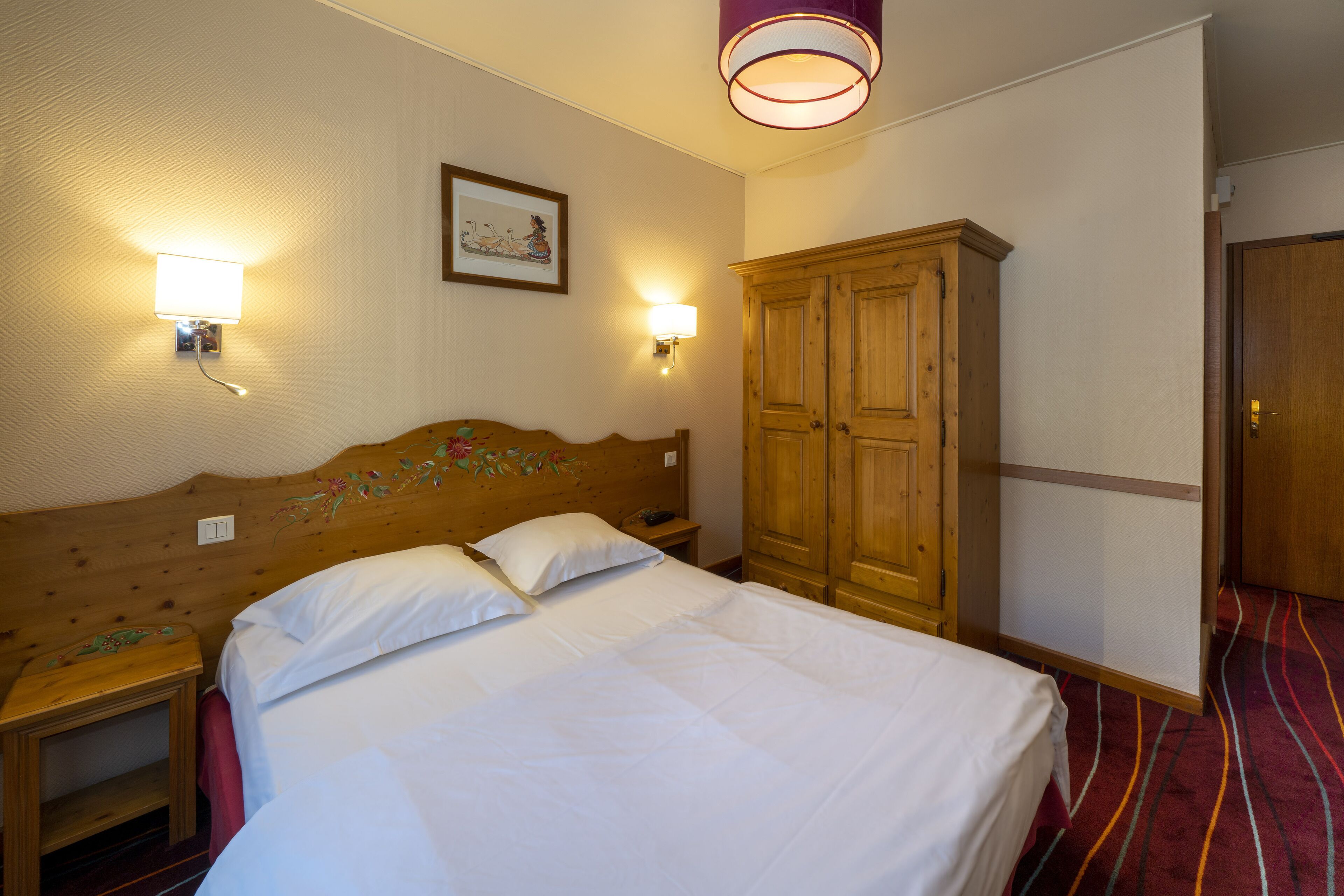 A hotel room with a wooden headboard, two bedside lamps, a wardrobe, and a bed with white linens.