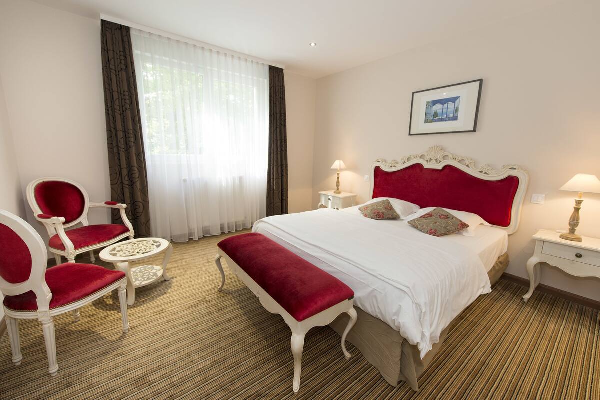 A hotel room with a large bed, two bedside tables, a red bench, two red chairs, and a small round table.