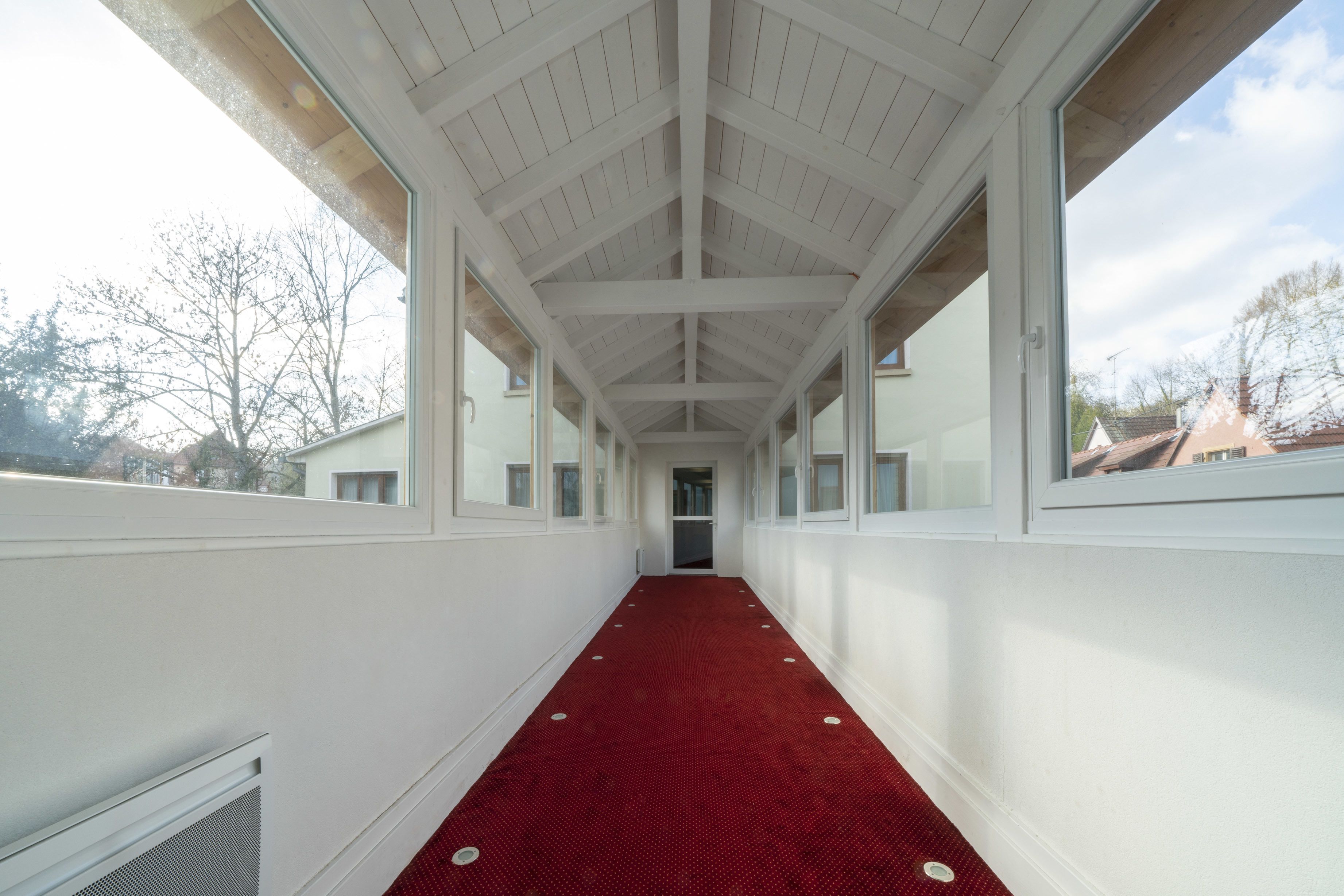A corridor with a red carpet, white walls, and large windows.