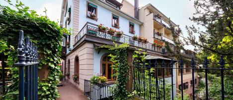 Un edificio de varios pisos con balcones decorados con flores, un sendero de piedra que conduce a la entrada y una cerca de metal que rodea la propiedad.