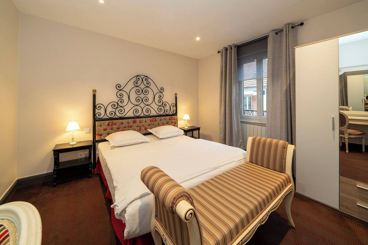 A hotel room with a large bed, a chair, a nightstand with a lamp, and a window with curtains.