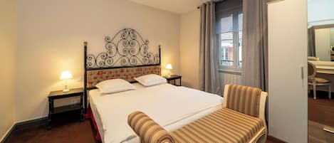 Habitación de hotel con una cama grande, una silla, una mesita de noche con lámpara y una ventana con cortinas.
