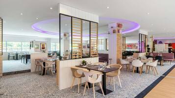 A modern hotel lobby with a central bar area, seating arrangements, and a view of the outdoor area.