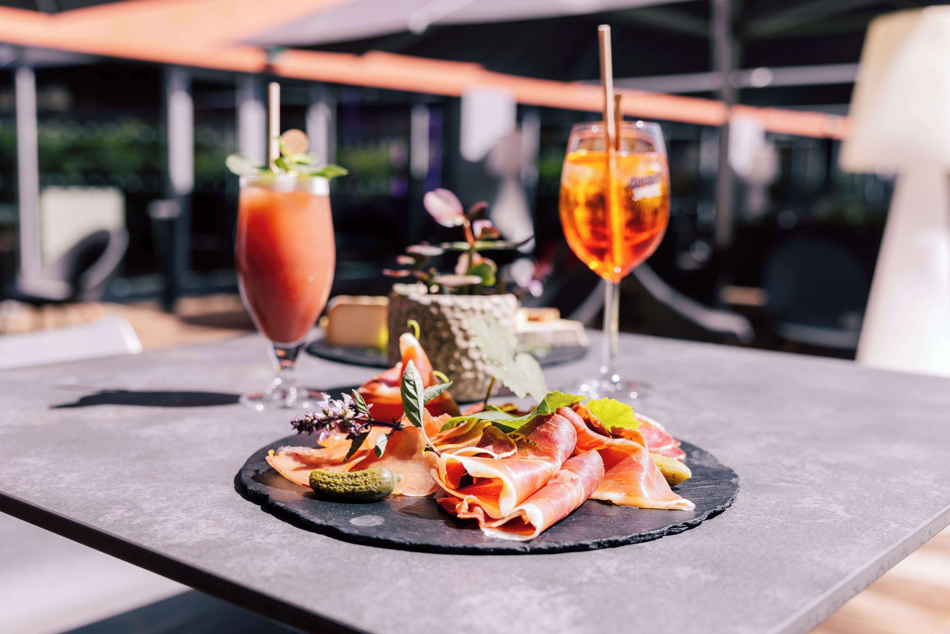 A table with a charcuterie board, a Bloody Mary, and a cocktail with a straw.