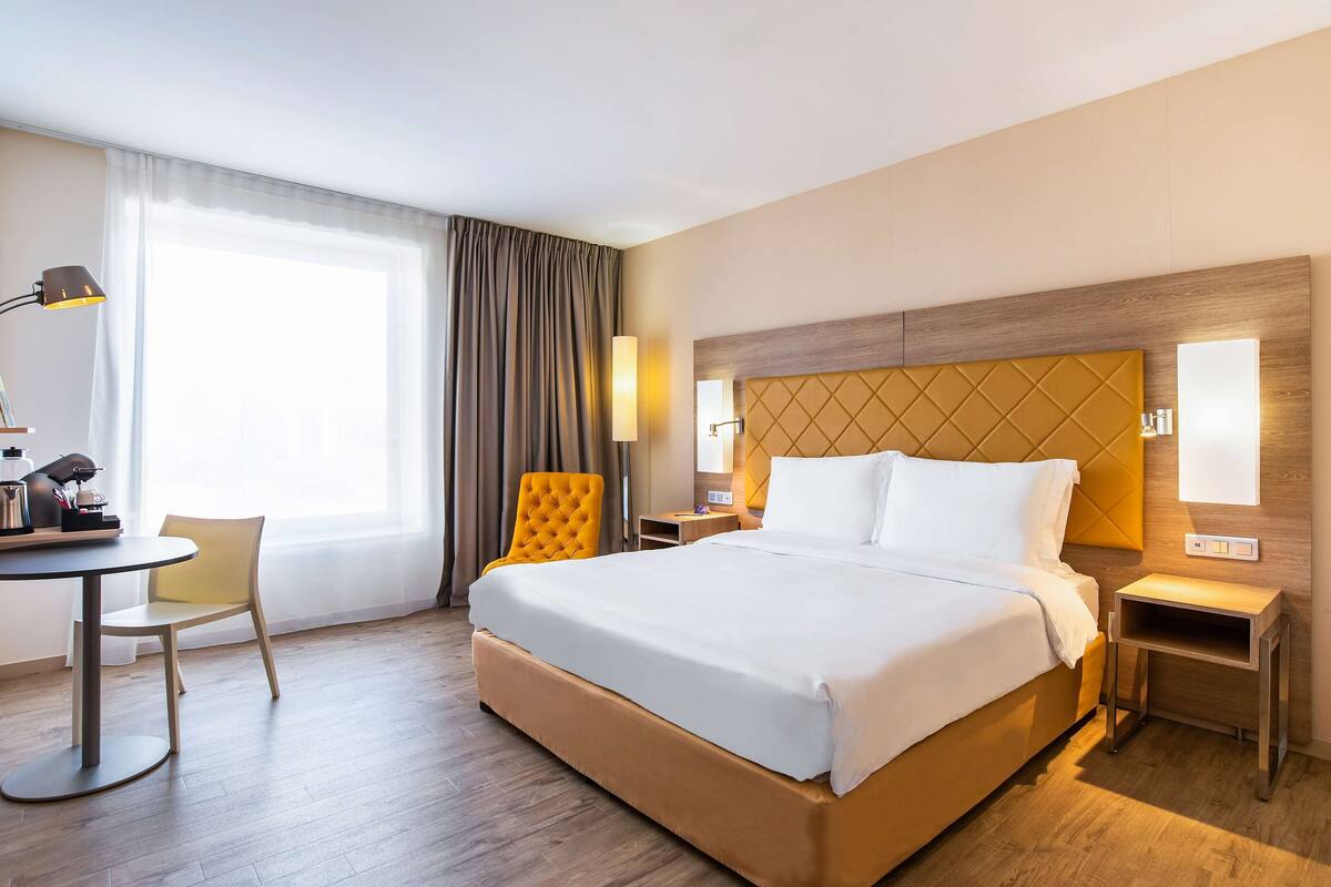 A hotel room with a large bed, a desk with a lamp, a chair, and a window with curtains.
