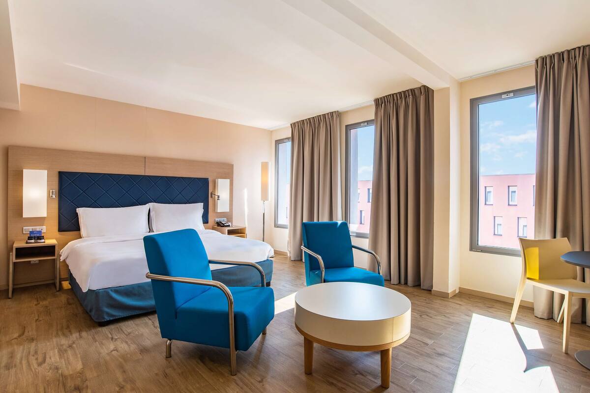 A hotel room with a bed, two blue armchairs, a round table, a nightstand, and a window with curtains.