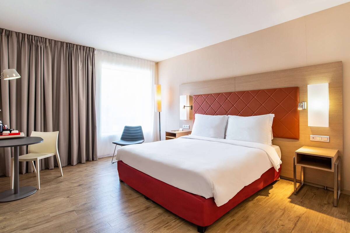 A hotel room with a large bed, a desk with a chair, a lamp, and a nightstand.
