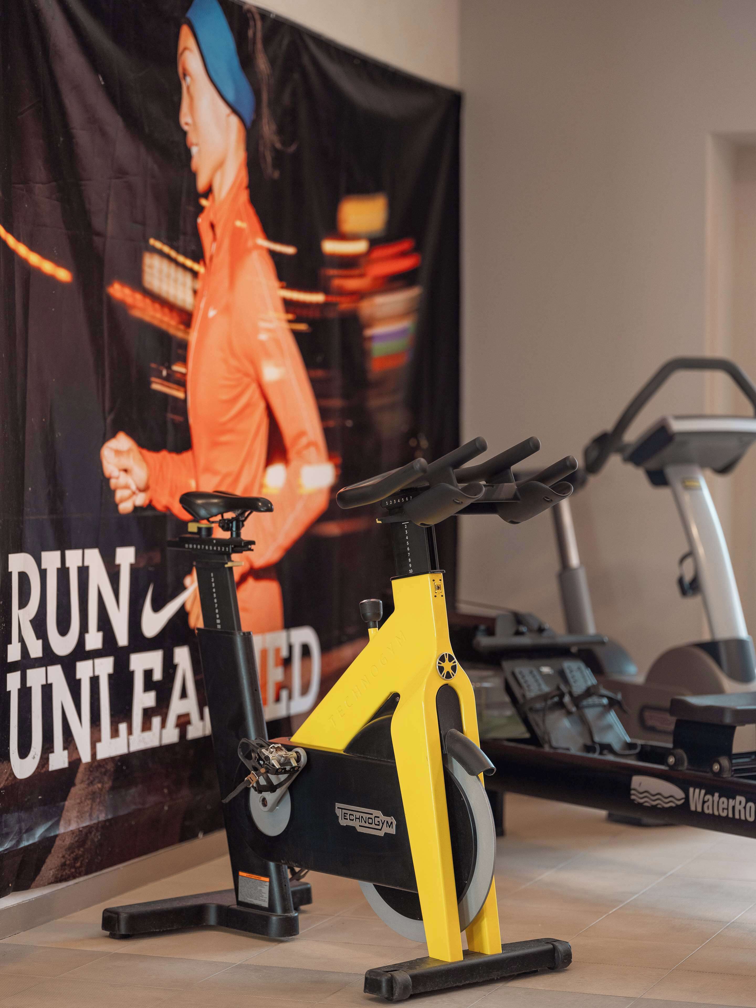 A stationary bike with a yellow frame and a water resistance feature, placed in a room with a treadmill and a motivational poster in the background.