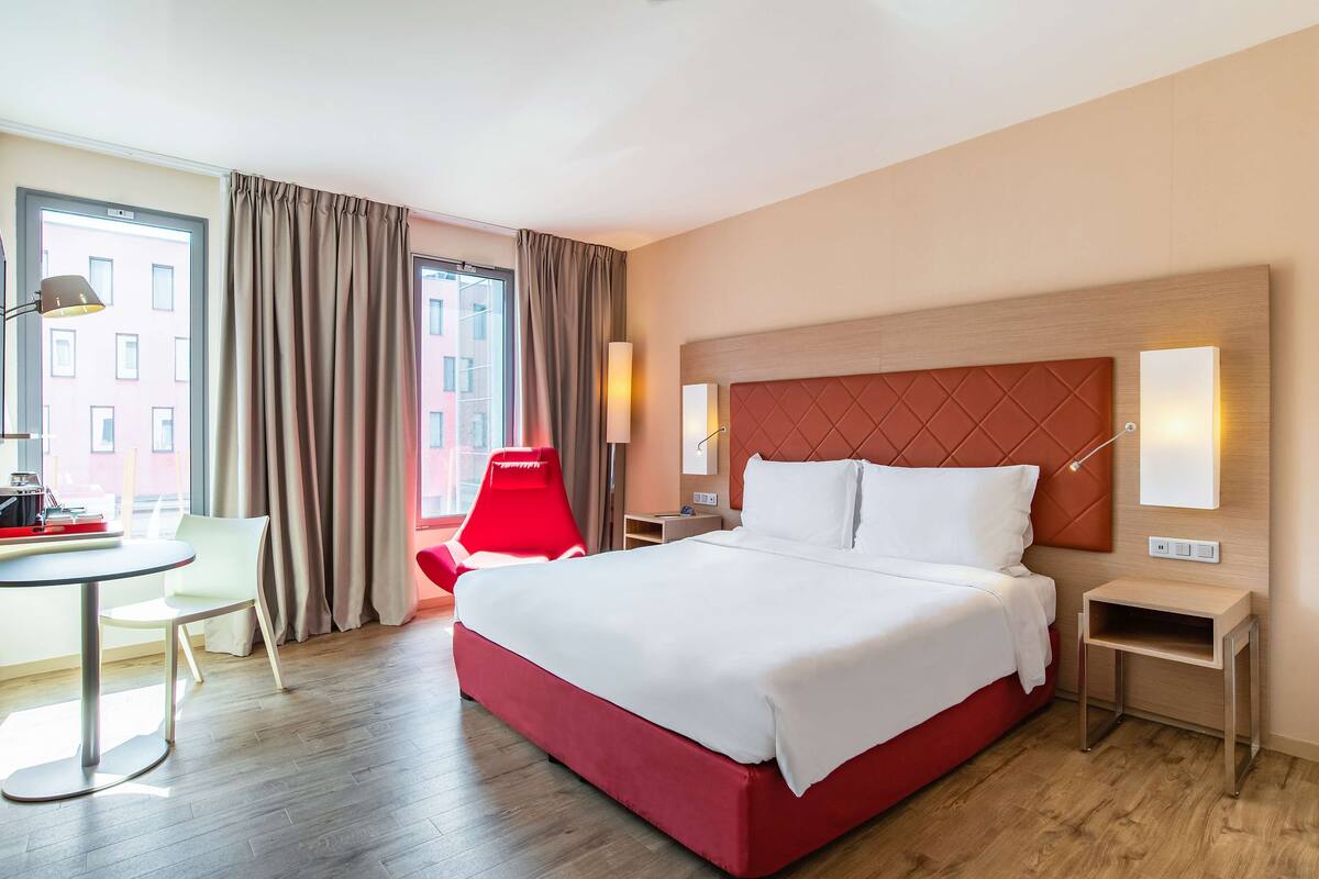 A hotel room with a large bed, a desk, a chair, and a view of the city.
