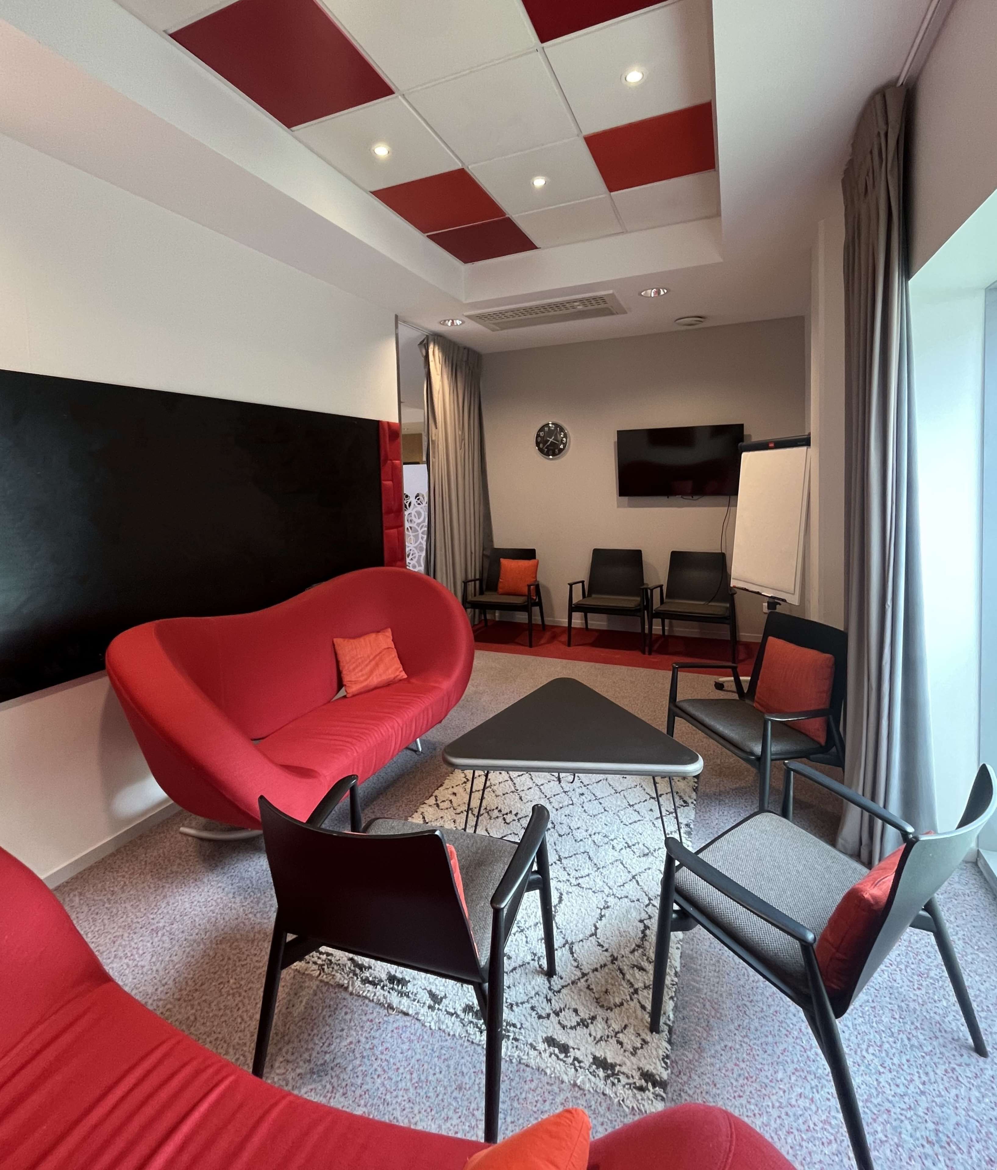 Una habitación moderna con sofá rojo, sillas negras y grises, alfombra blanco y negro, televisor de pantalla plana y un ventanal con cortinas.