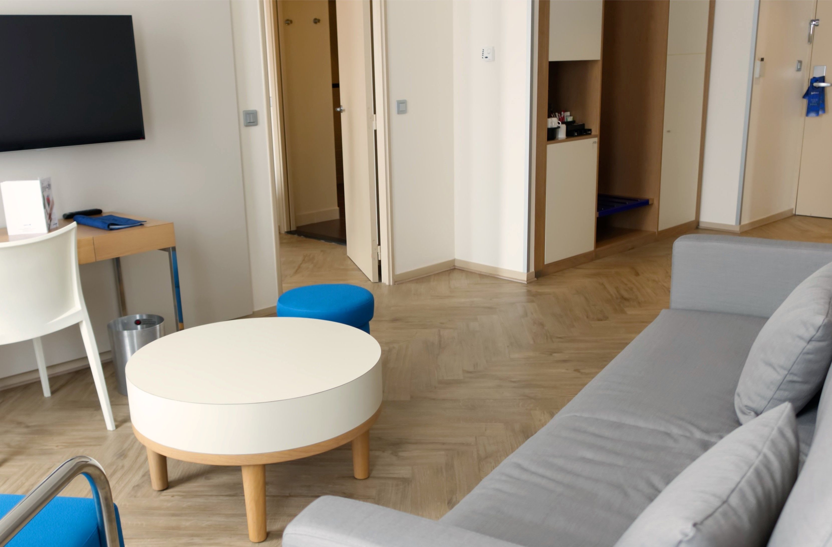 Un salon moderne avec une table ronde blanche, un canapé gris et un pouf bleu. Un téléviseur est fixé au mur, et il y a un petit bureau avec une chaise.