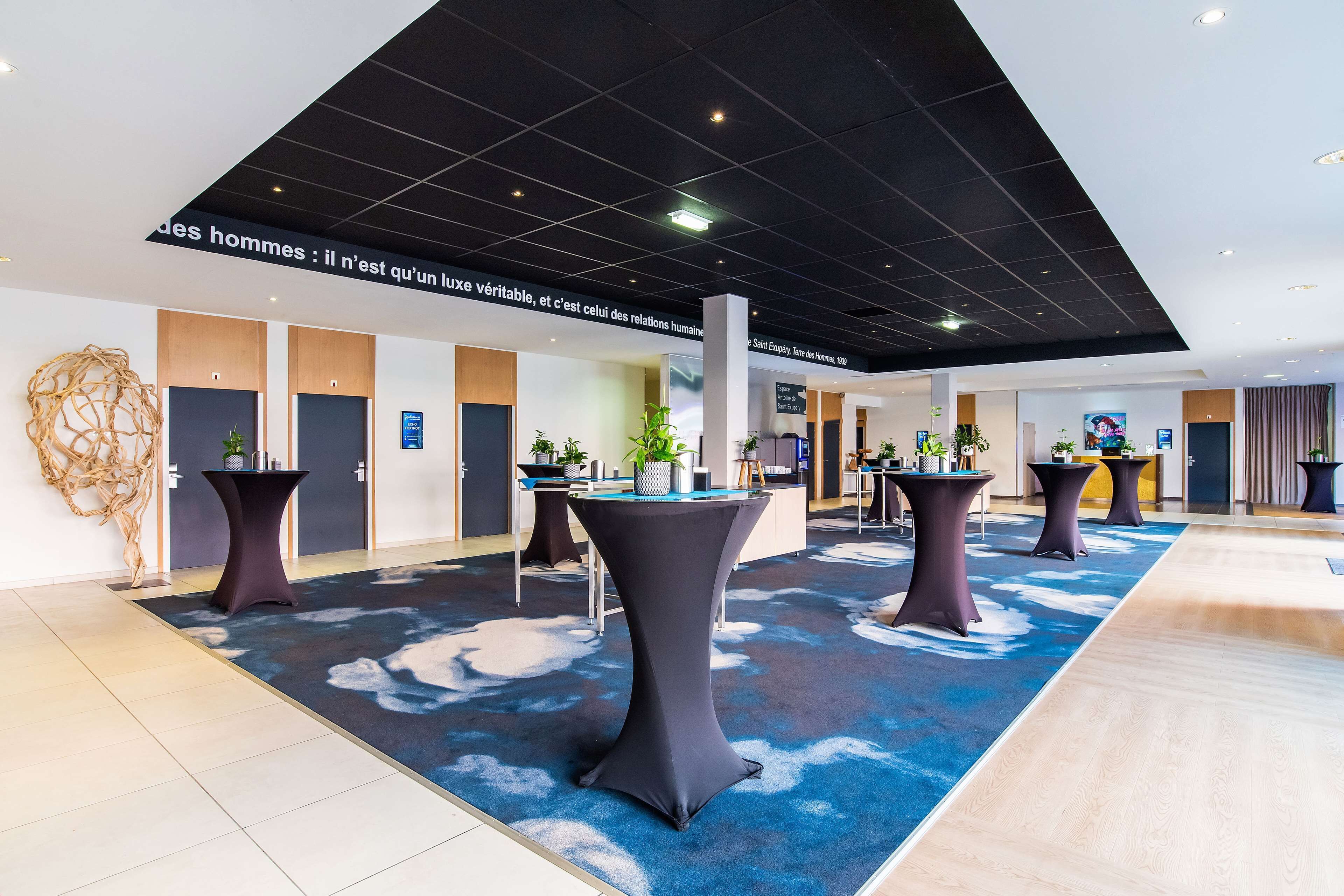 En modern hotellobby med högt i tak, infälld belysning och ett blått golv med molnmönster. Det finns flera runda bord med mörka dukar och krukväxter.