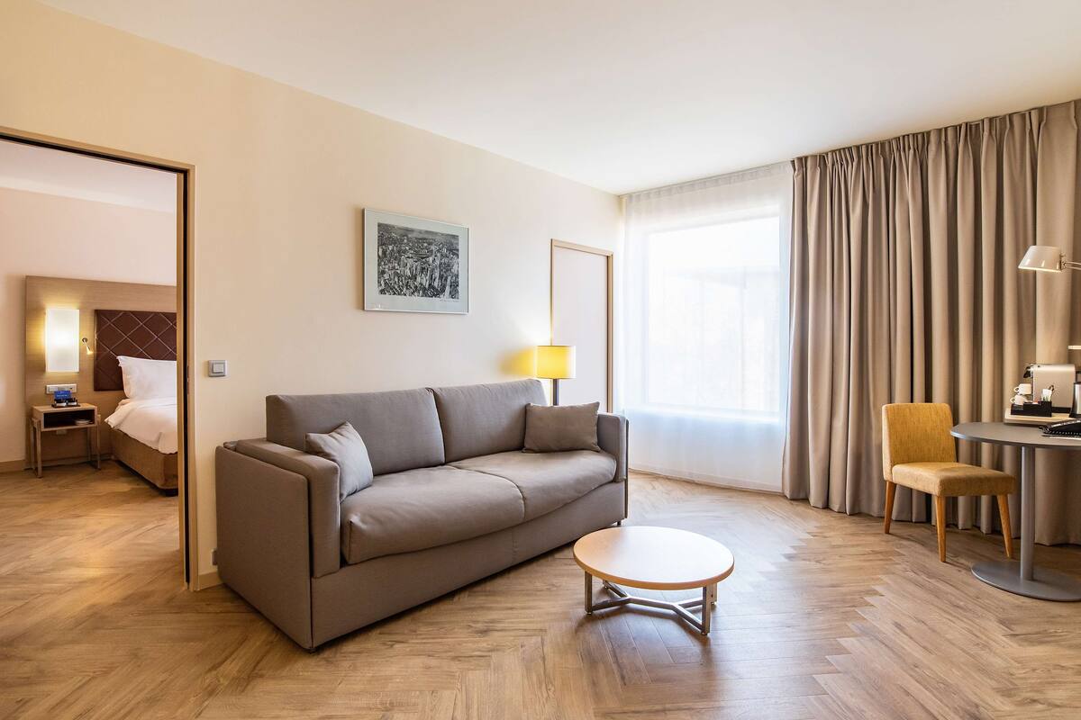 A modern hotel room with a sofa, a small round table, a lamp, a chair, and a bed in the background.
