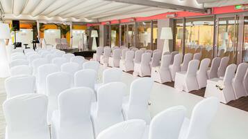 Une salle de banquet avec des rangées de chaises et de tables blanches, une scène et un espace de réception comprenant un comptoir et des chaises.