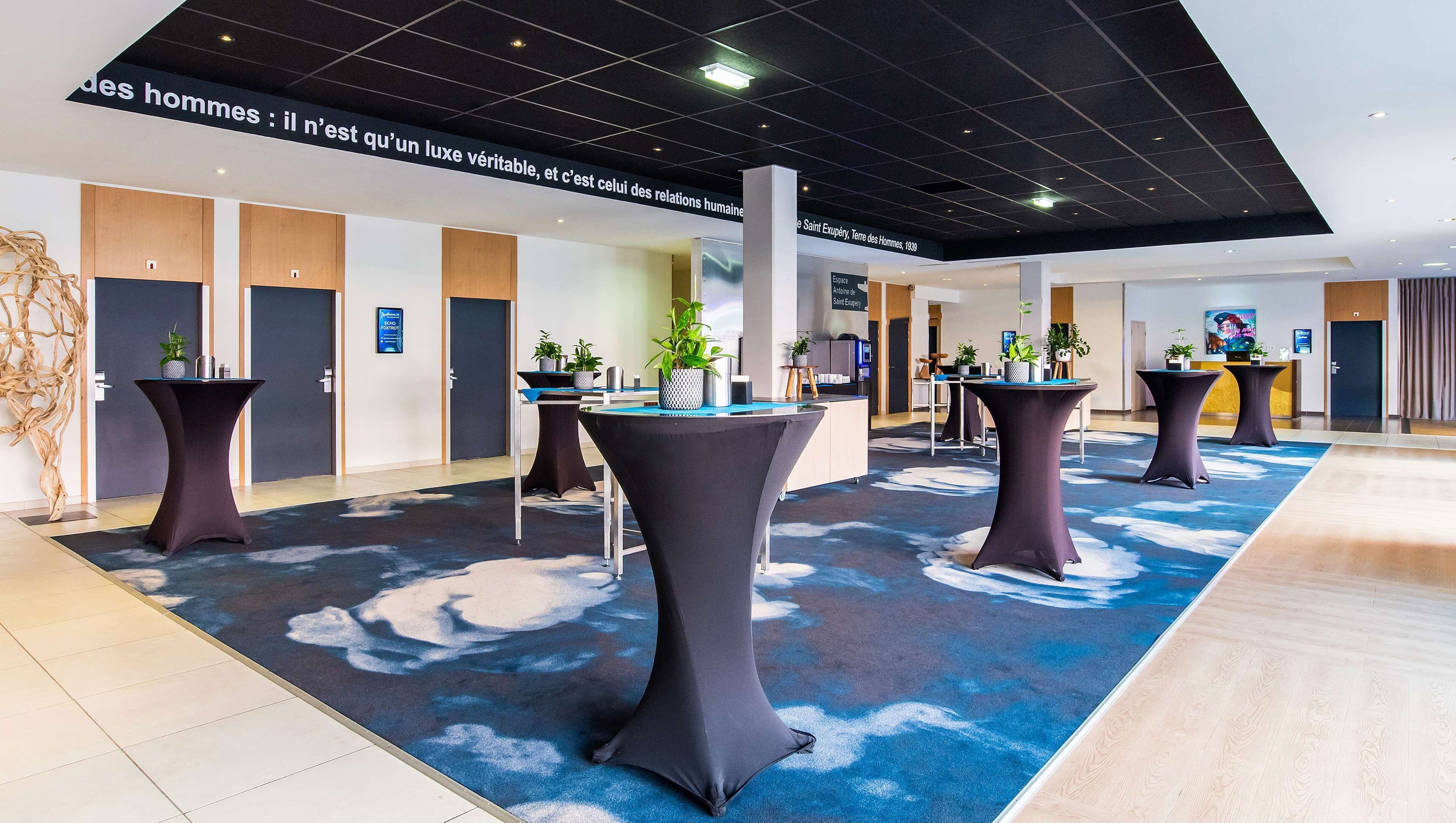 En modern hotellobby med högt i tak, mörka takpaneler och en blå matta med molnmotiv. Det finns flera runda bord med mörka dukar, och varje bord är omgivet av krukväxter. Väggarna är dekorerade med inramade konstverk, och det finns trädörrar längs den bakre väggen.