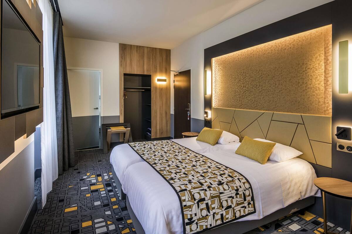 A modern hotel room with a large bed, a built-in wardrobe, a small table, and a wall-mounted light fixture.