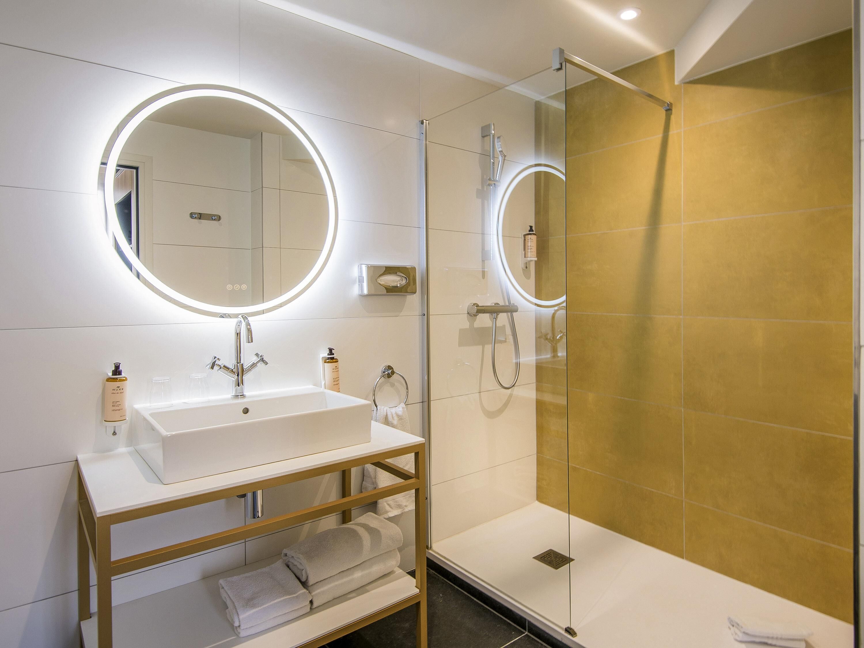 A modern bathroom with a white sink, a mirror with a circular light, a glass shower enclosure, and a gold-tiled shower wall.