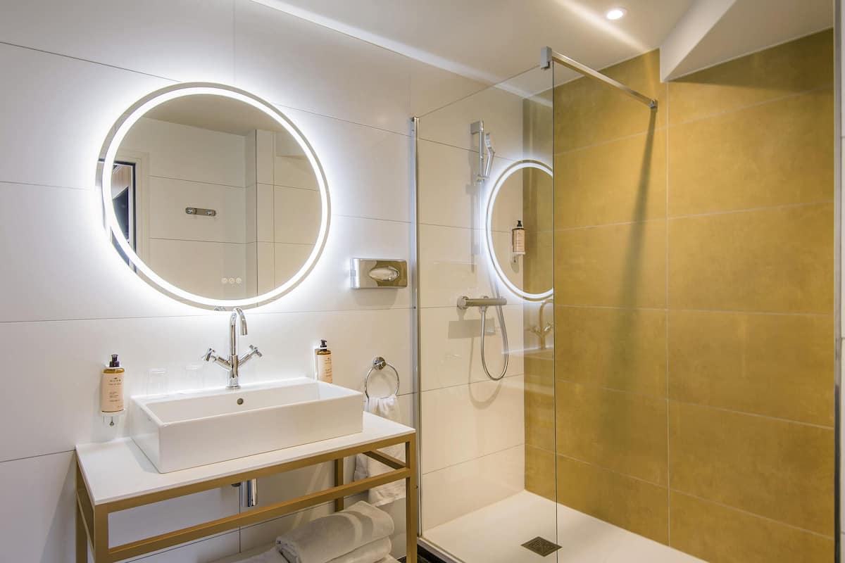 A modern bathroom with a white sink, a mirror with a circular light, a glass shower enclosure, and a gold-tiled shower wall.