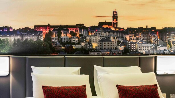 Habitación de hotel con vista a la ciudad al atardecer, que incluye una cama grande con ropa blanca y almohadas rojas estampadas, una mesita de noche pequeña y un buró.
