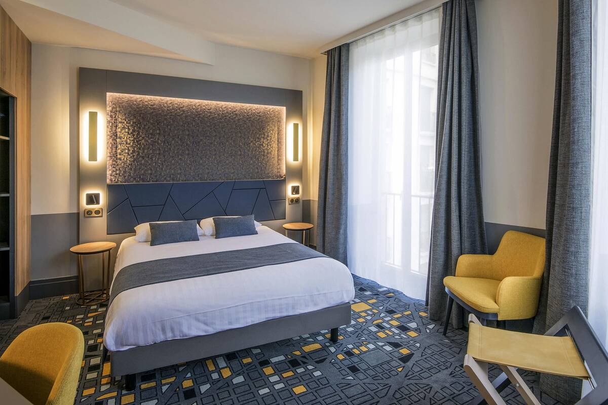 A modern hotel room with a large bed, a headboard with integrated lighting, a small side table, a chair, and a view of the city through large windows.