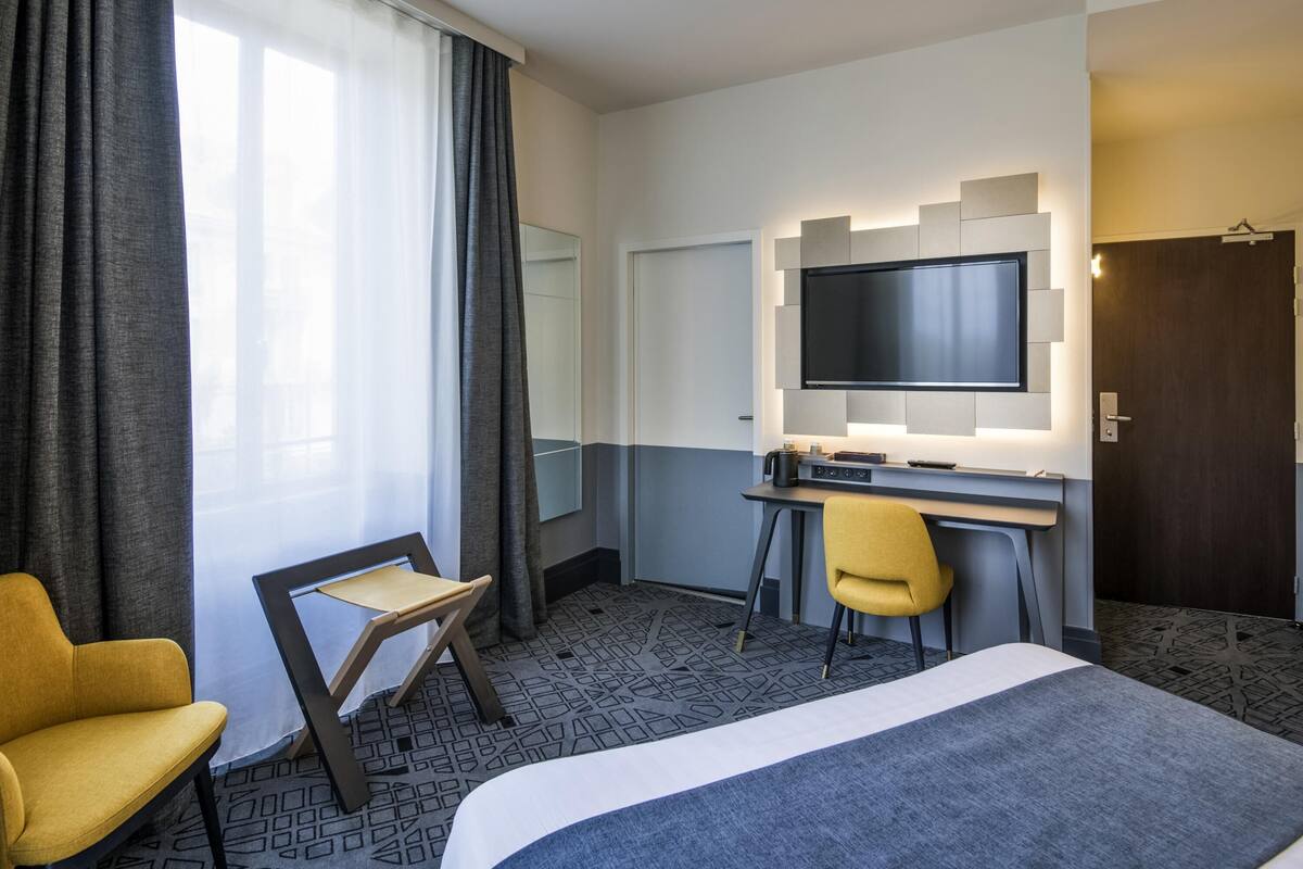A hotel room with a bed, a desk, a TV, a chair, and a window with curtains.