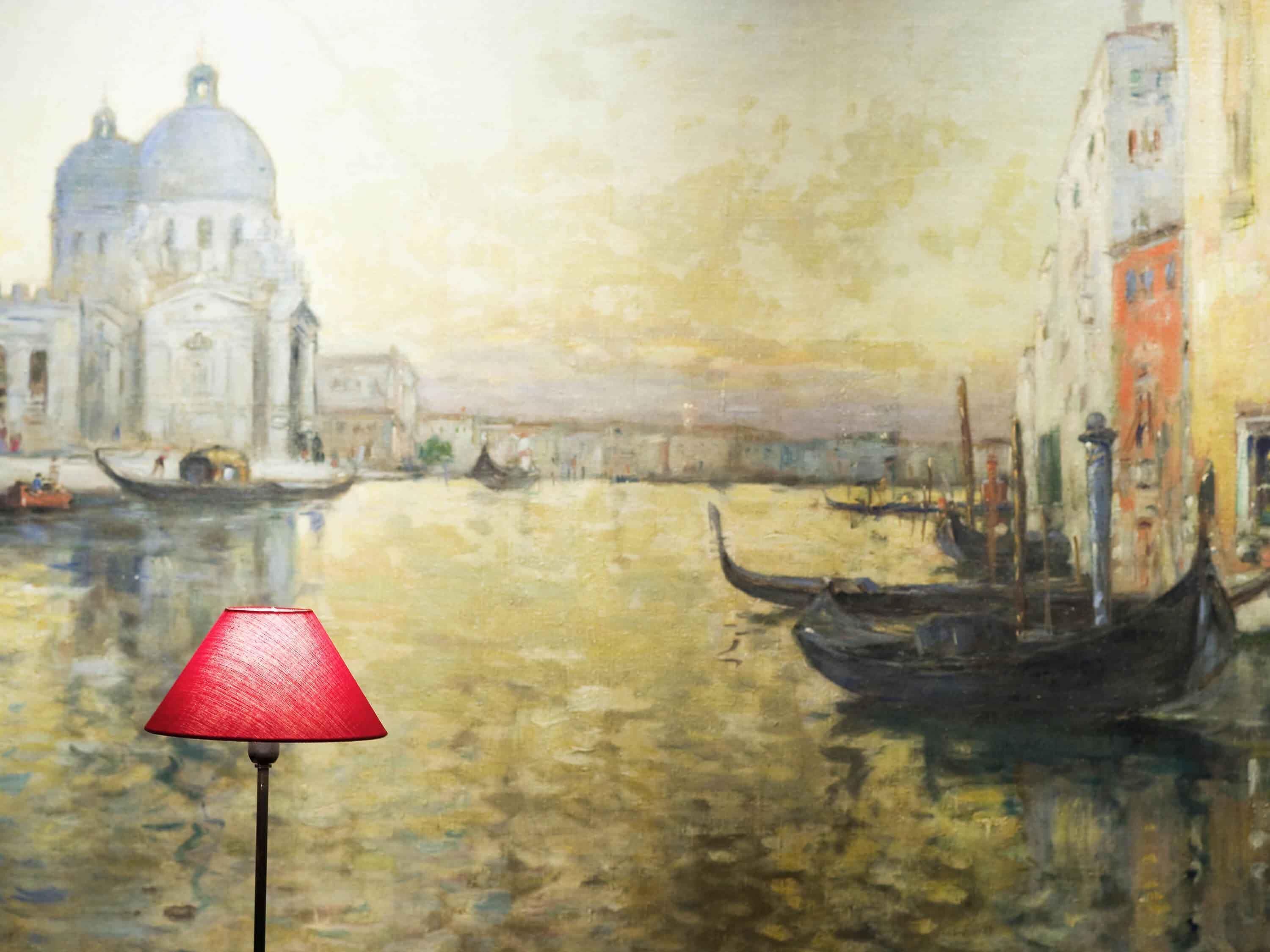 Una pintura de un canal con góndolas y edificios, con una lámpara roja en primer plano.