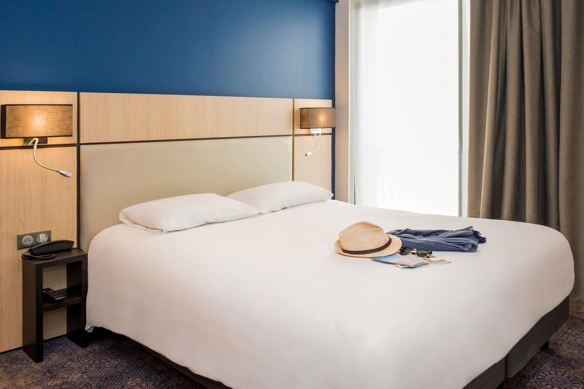 A hotel room with a bed, bedside lamps, a nightstand, a hat, and a folded towel on the bed.