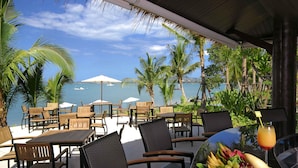 A seaside patio with wooden tables and chairs, umbrellas, and a table with fruit and a drink. - ibis Samui Bophut (Koh Samui)