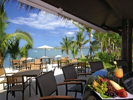 Un patio frente al mar con mesas y sillas de madera, sombrillas y una mesa con fruta y una bebida.