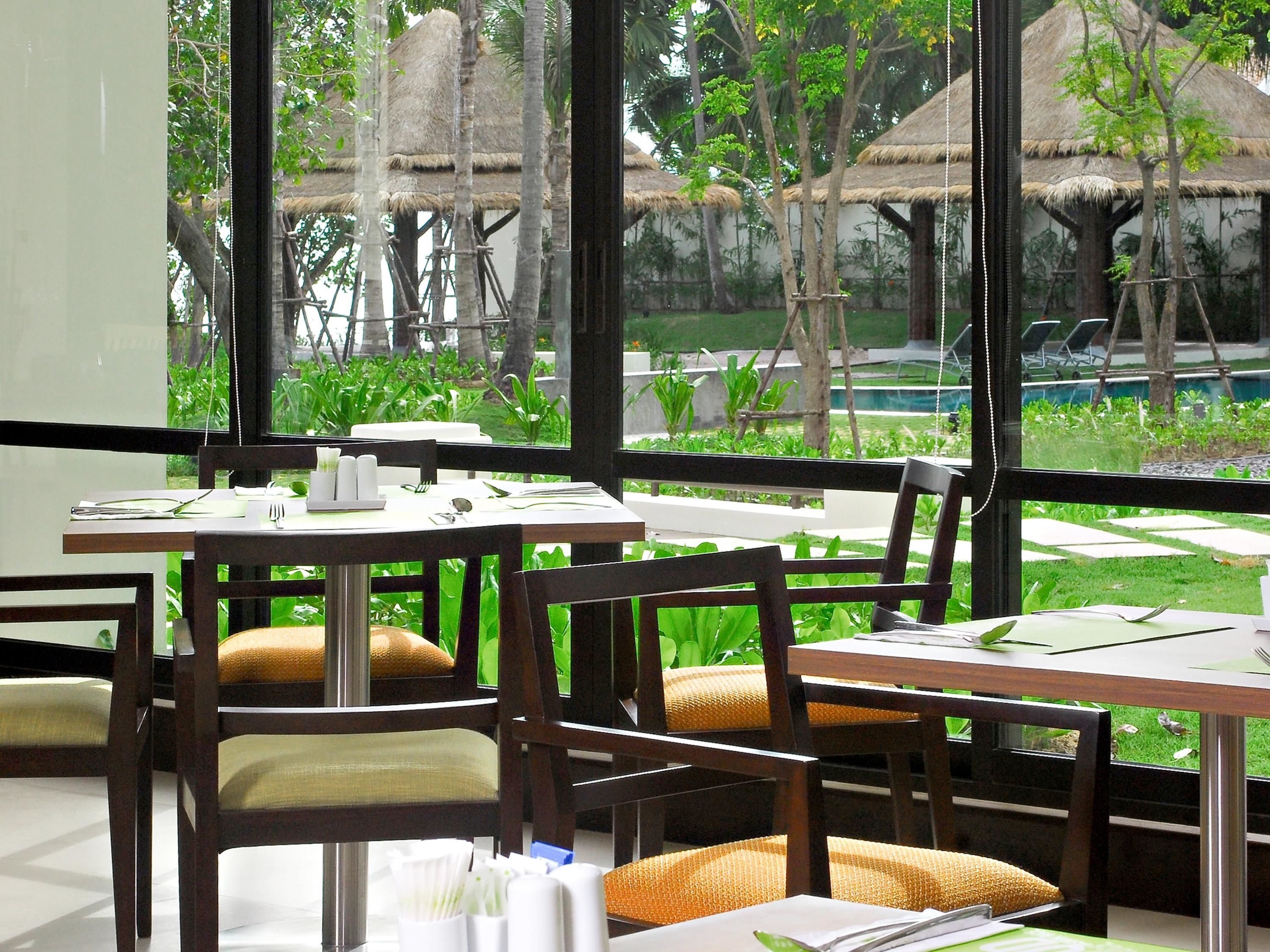 A restaurant with tables and chairs, a view of a pool and garden through large windows.