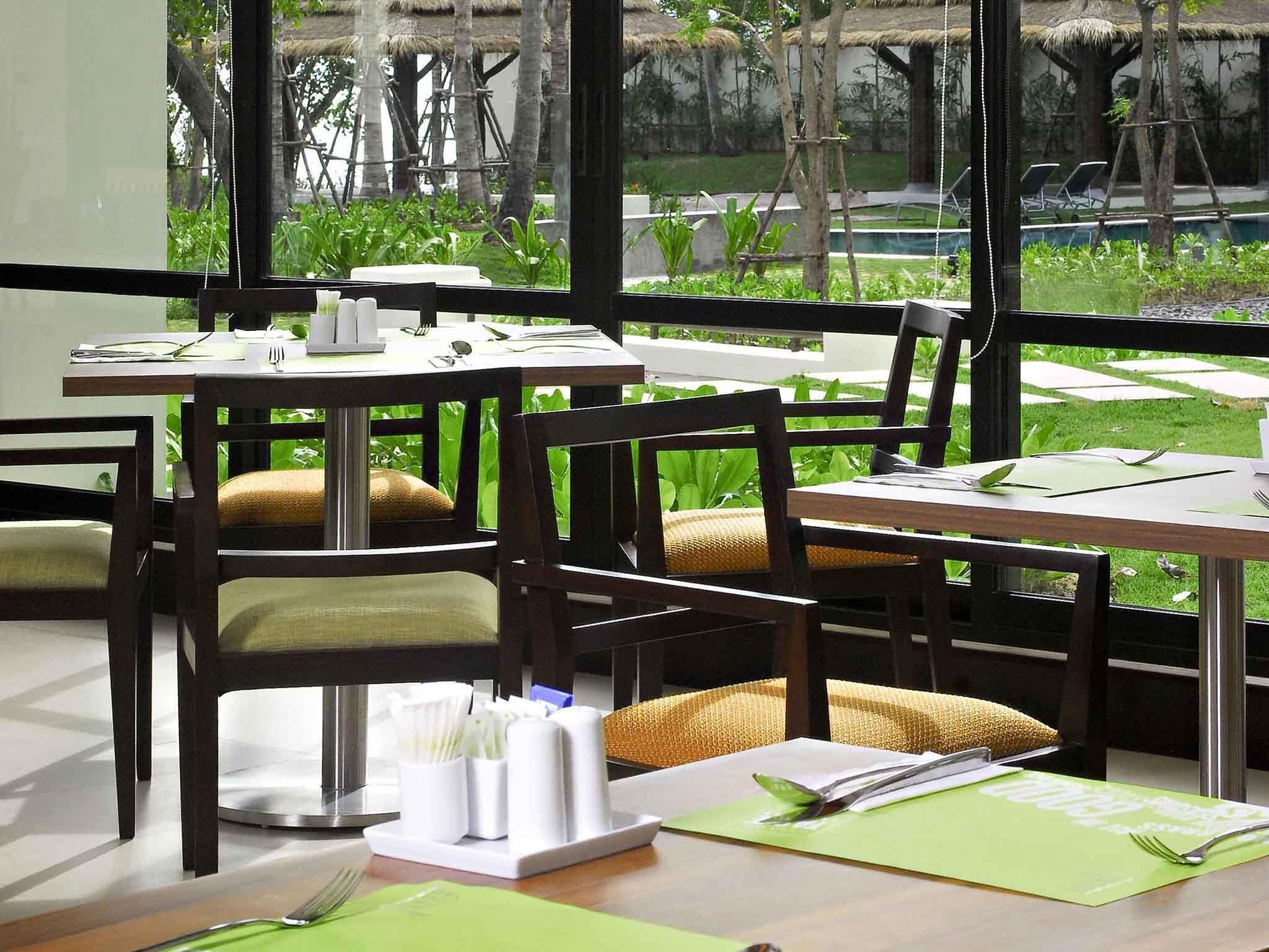 A dining area with tables and chairs, a view of greenery outside, and a pool in the background.